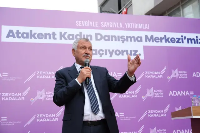 Karalar resmi giyimli mor bir platformun önünde elinde mikrofon var ayakta. 