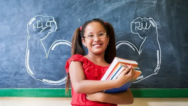 Niña en clase frente a un pizarrón en el que están dibujados a cada lado de ella brazos musulosos