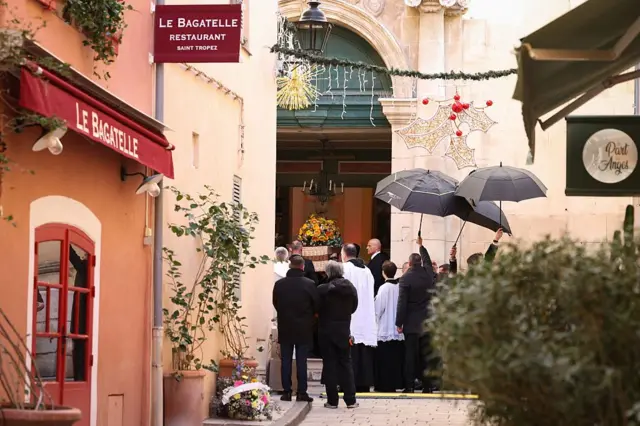 Улица, с которой виден вход в церковь. Видно, что из дверей выносят гроб. Стоящие у дверей люди подняли вверх три черных зонта.