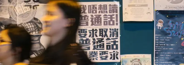 香港浸会大学逸夫校园内一群女学生走过贴上了反对普通话毕业门槛豁免试标语的学生会民主墙(BBC中文网图片22/1/2018)