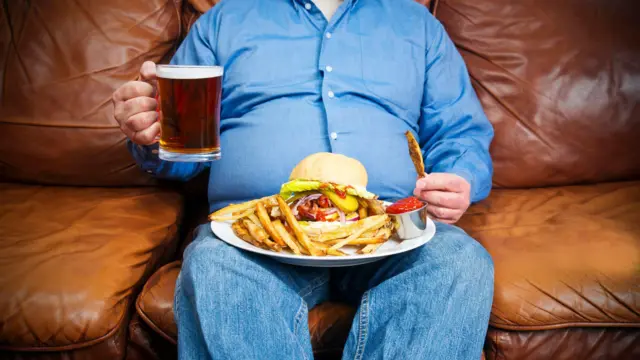 homem corpulento, vestindo uma camisa social azul e calça jeans, segura um prato com hambúrguer e batatas fritas no colo, enquanto mantém uma caneca de cerveja na mão