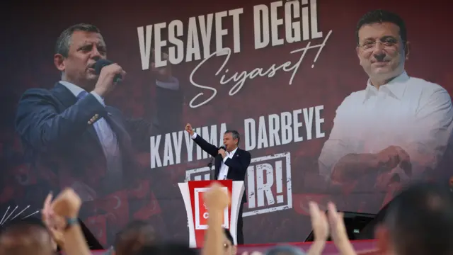 CHP genel başkanı özgür özel bir mitingde kürsüde, arkasında ekrem imamoğlu'nun fotoğrafı var, üzerinde "kayyıma, darbeye hayır" ve "vesayet değil, siyaset" mesajları yazılı. önde mitingi izleyen kalabalığın elleri görülüyor. 14 Eylül 2025, Ankara