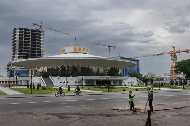 Здание в форме «летающей тарелки» в центре Бишкека. На крыше крупными буквами написано «Цирк». На заднем плане — три строительных крана и строящаяся высотка.