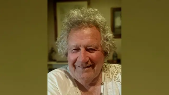 Andrew St. Helier Bourke tiene el pelo canoso y sonríe a la cámara. Lleva una camiseta blanca con un estampado verde. El fondo está desenfocado. Hay cuadros enmarcados en la pared detrás de él.