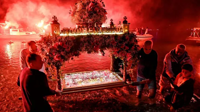 Nekoliko muškaraca nosi ukrašeno drveno postoje kroz vodu dok je iza njih vatromet