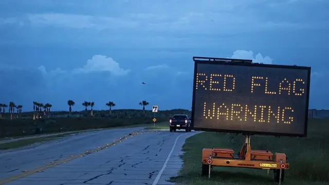 Foto de uma estrada, com um carro no fundo e no primeiro plano uma placa luminosa escrito em inglês 'aviso de bandeira vermelha'.