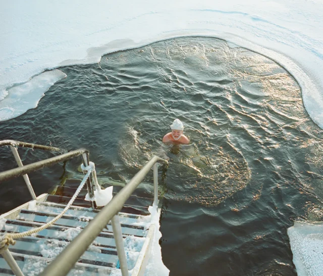 Starszy mężczyzna w białej czapce na głowie kąpie się w przerębli. Na pierwszym planie widać pokryte lodem schody prowadzące do wody. 