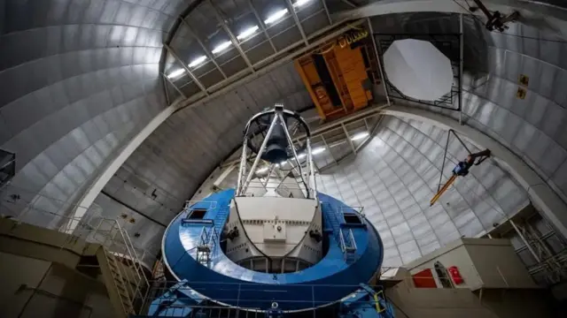 На фотографии показано внутреннее пространство огромного металлического купола с изогнутыми серебристыми панелями, возвышающимися над головой, как внутреннее пространство гигантской стальной оболочки. В центре стоит массивная сине-белая телескопическая установка в форме толстого кольца с высокой открытой рамой над ней, на которой установлены главный зеркало и приборы. К конструкции прикреплены проходы, перила и лестницы, а под потолком висит оранжевый подвесной кран, придающий сцене индустриальный, соборный вид.