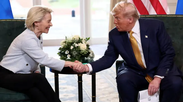 Von der Leyen, de jaqueta branca curta e calça preta, estende a mão diante de uma mesa lateral com um arranjo de flores brancas para cumprimentar Trump, que usa terno azul, gravata dourada e segura papéis na outra mão.