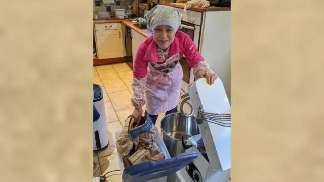 三角巾にエプロン姿の日本人女性が、バケツの中の小麦粉をパンをこねる機械に移そうとしている