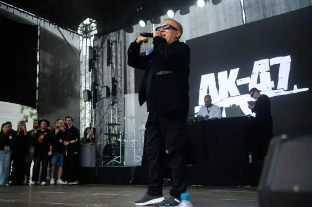 Мужчина в солнечных очках с микрофоном на фоне экрана с надписью АК-47