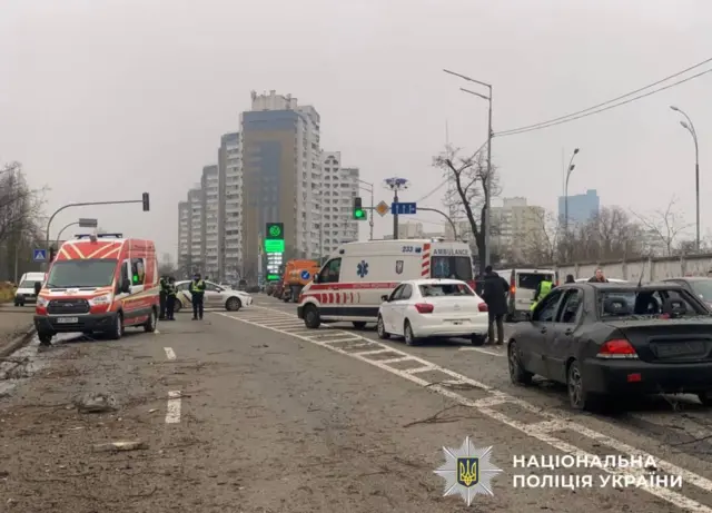 Всіяна уламками дорога, на якій стоять пошкоджені авто. Вдалині видніється висотка 