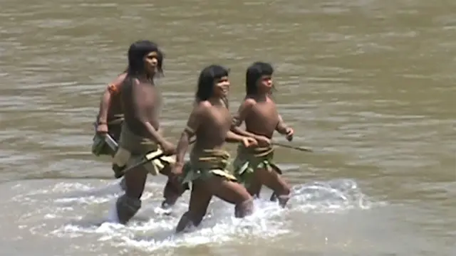 Três mulheres Mashco Piro caminham pela água rasa de um rio. Usam saias feitas de materiais naturais, como folhas ou fibras vegetais, e carregam longas lanças de madeira.