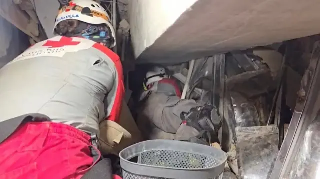 コンクリートの大きいかたまりや金属製の建物の一部がひしゃげて多い重なっている。その隙間にできたわずかな空間に、灰色に赤十字に着いた隊服の救助隊員が腹ばいになって潜り込もうとしているのを、手前で同僚の隊員がしゃがんで見守っている