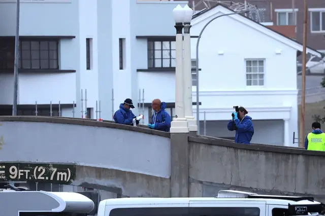 Mavi kıyafetli adli tıp ekipleri köprünün üzerinde çalışma yürütüyor. Eldivenli bir kişi fotoğraf çekiyor.