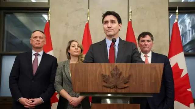 Kanada Başbakanı Justin Trudeau kürsüde konuşuyor, arka planda diğer yetkililer ve Kanada bayrakları görülüyor