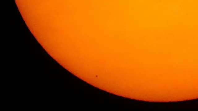 A imagem mostra um pequeno ponto preto na parte inferior de uma imensa bola laranja que é o sol.