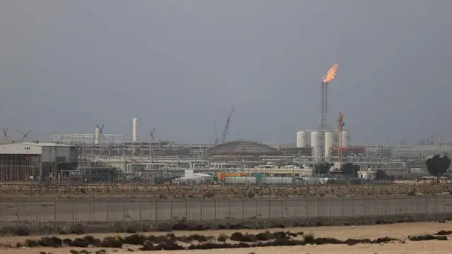 灰色の空を背にしたエネルギー施設で、高いフレアスタックの煙突から炎が出ている。巨大な施設の手前に柵と、砂地が広がる