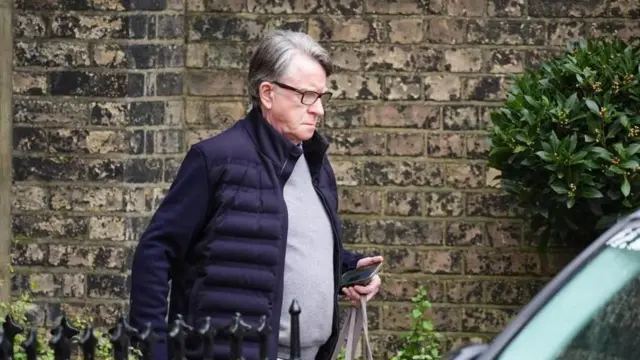 ベージュ色の石造りの壁を背に、黒い手すりの向こうから外へ出る白髪交じりで眼鏡の高齢男性。紺色のジャケット、灰色のセーターを着て、手にトートバッグとスマートフォンを持っている。手前に警察の覆面車が停まっている