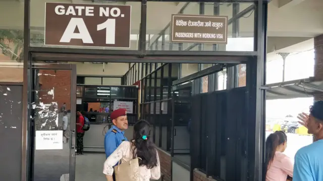 त्रिभुवन अन्तर्राष्ट्रिय विमानस्थलमा वैदेशिक रोजगारीमा जाने यात्रुहरूका लागि छुट्ट्याइएको प्रवेशद्वारअगाडि उभिएका प्रहरीसँग कुरा गर्दै एक महिला, बाहिर अर्का एक पुरुष र एक महिला देखिन्छन्