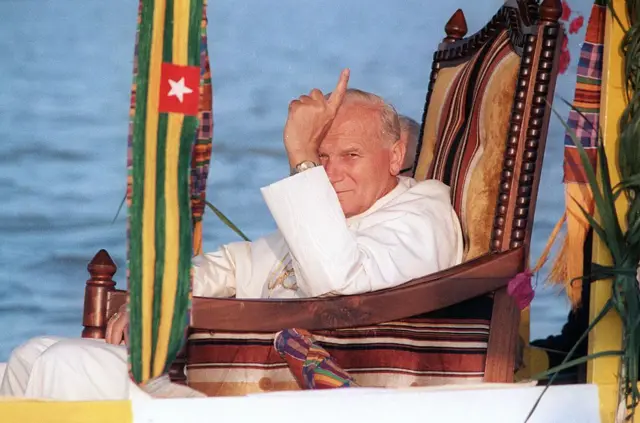 Le Pape Jean-Paul II assis dans un fauteuil transporté dans une pirogue sur le lac Togo lors de sa visite à Togoville en 1985.