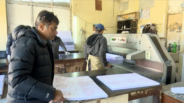 मतपत्र छपाई चल्दाको क्षण। एक जना कामदार मशिनतर्फ उभिएर काम गरिरहेका छन् भने कालो ज्याकेट लगाएका अर्का व्यक्ति छापिएको मतपत्र हेरिरहेका छन्।