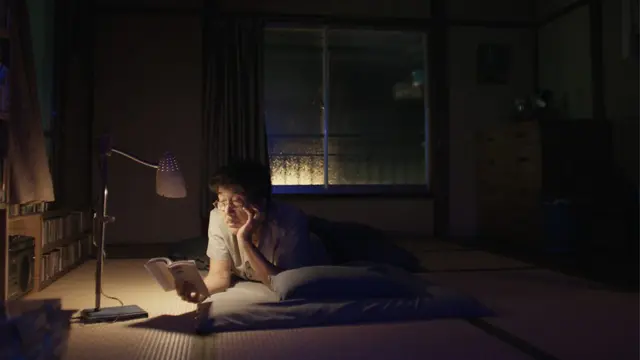 O ator Koji Yakusho lê um livro sob a luz de um abajur, deitado de bruços numa cama sobre um tatame, em cena do filme Dias Perfeitos