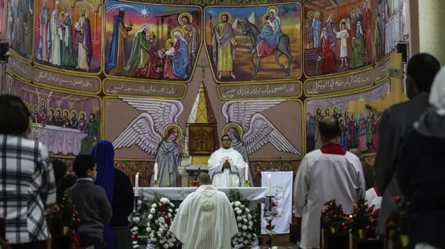 A priest holds a Christmas Eve ceremony while people pray in front of a brightly decorated wall depicting Chritsian scenes inside the Holy Family Catholic Church in Gaza City, on 24 December 2018