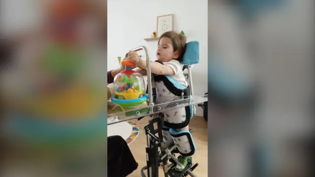 Laia est attachée à une structure métallique qui lui permet de se tenir debout et de jouer avec ses jouets.