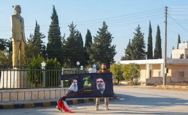مقاتل يحمل صورة ممزقة لزعيم حزب الله الراحل السيد حسن نصر الله وهو يقف بالقرب من تمثال الرئيس السابق حافظ الأسد في نبل شمال حلب بسوريا في 2 ديسمبر 2024.
