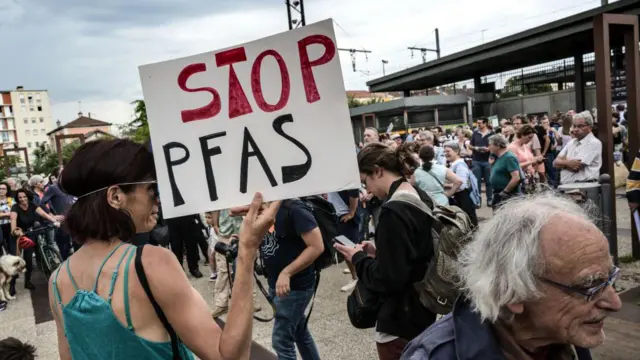 क्यामेरापट्टि आफ्नो ढाड फर्काएर भिडमा उभिएकी एक महिलाले बोकेको एक सङ्केतमा "स्टप पीएफएएस" भनेर लेखिएको छ। कालो कपाल लगाएकी उनले नीलो लुगा लगाएकी छन् 