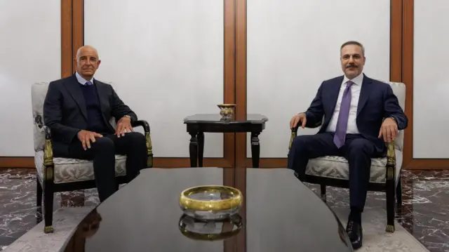 İki adam yan yana kaltuklarda oturmuş. Takım elbise giymişler. GAzetecilere poz veriyorlar.