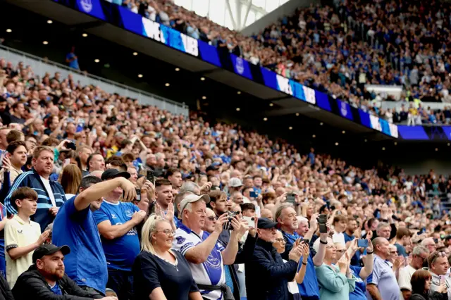 Sute de fani Everton, îmbrăcați în albastru și alb, stau în Hill Dickinson Stadium și își ridică telefoanele în timpul unui meci amical dintre Everton și AS Roma în 2025.