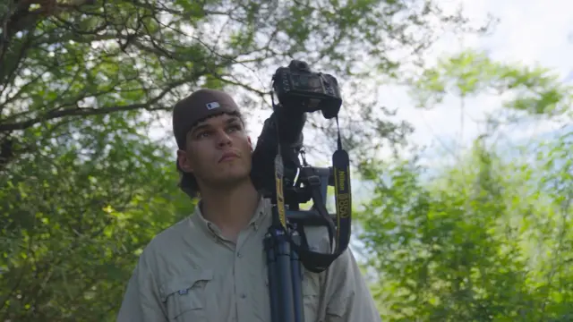 A young man standing outdoors in a wooded area, carrying a large camera with a telephoto lens mounted on a tripod over his shoulder.