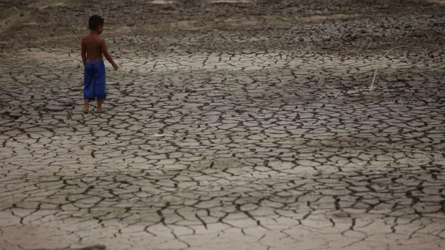 Um menino, sem camisa e de bermuda azul, caminha sobre um chão seco, cheio de rachaduras