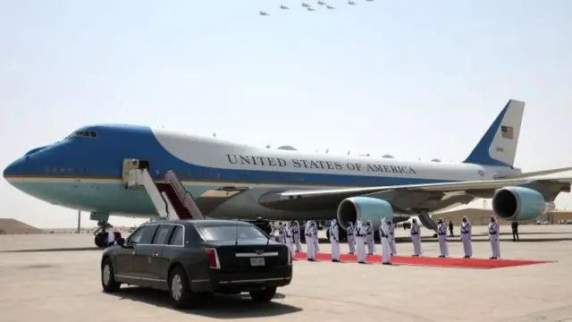 El Air Force One azul y blanco está en la pista de un aeródromo de Qatar. Funcionarios vestidos de blanco se sitúan a ambos lados de una alfombra roja que sale de la puerta del avión. 