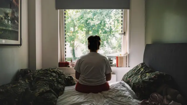 একজন নারী বিছানায় বসে জানালা দিয়ে বাইরের দিকে তাকিয়ে আছেন 