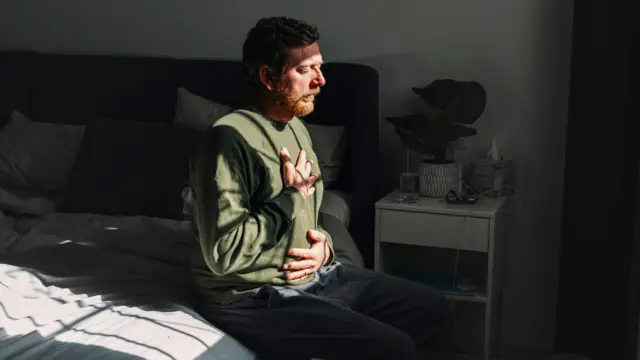 一个男人坐在床边，一只手放在胸口，另一只手放在腹部，窗外的阳光洒在他的脸上。