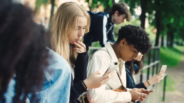 A group of teenagers look at their phones while leaning against railings. In focus is a blonde girl and a boy with dark curly hair.