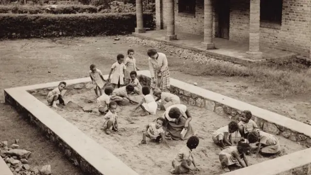 Um grupo de crianças brincando em uma caixa de areia. Há duas mulheres com elas. A foto é em preto e branco