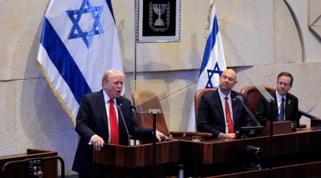 Trump kürsüde konuşuyor, yanında iki takım elbiseli siyasetçi onu dinliyor