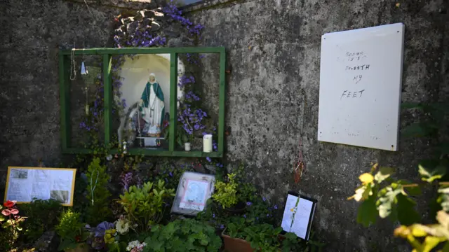 Dos altos muros de piedra se cruzan en ángulo recto en un jardín. En la esquina del ángulo se encuentra una gruta centrada en una estatua de la Virgen María. Está rodeada de flores y arbustos. Hay velas en un estante cerca de los pies de la estatua. También hay mensajes debajo de la estatua dejados por la gente y, a la derecha, una pizarra blanca con un mensaje de recuerdo.
