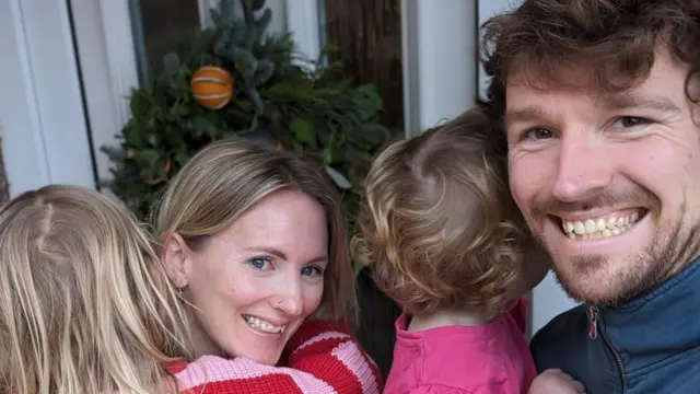 Louise y Pete sonríen a la cámara, sosteniendo a sus dos hijas.