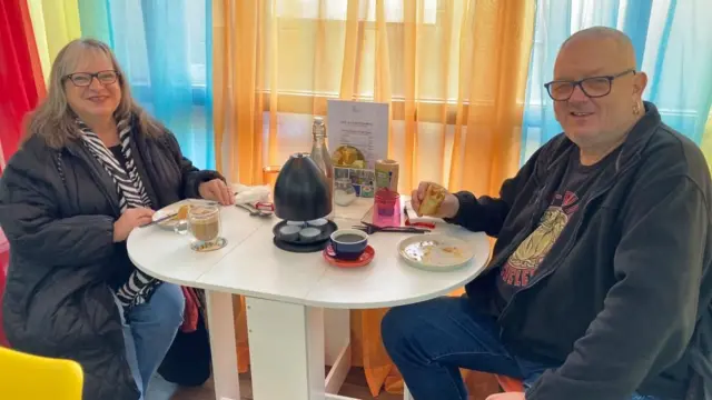 Linda, sentada a la izquierda de una pequeña mesa ovalada, sonríe y mira a la cámara, con comida y un café delante. Su esposo, Peter, está a la derecha, sosteniendo un panecillo de salchicha y mirando a la cámara. Linda lleva vaqueros, un abrigo negro, una bufanda blanca y negra alrededor del cuello, cabello largo y gafas. Peter lleva gafas y ropa oscura. Lleva un pendiente en la oreja izquierda.