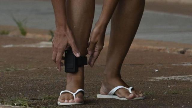 Detento com mãos próximas a tornozeleira eletrônica instalada no tornozelo direito dele