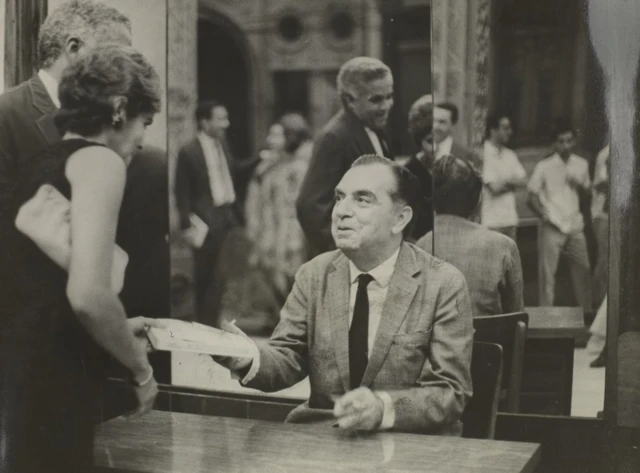 Rodrigues sorrindo diante de mesa, entregando livro para pessoa, aparentemente em sessão de autógrafos; há outras pessoas à sua volta