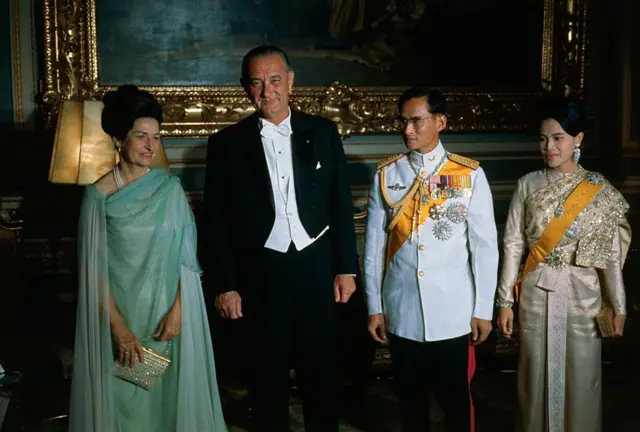 Королевская чета вместе с президентом США и его женой. Король - в военном мундире, Сирикит - в богато украшенном серебрянном платье, Джонсон - в смокинге, а его супруга - в свободном зеленом платье