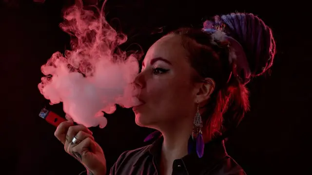Une femme, en demi-silhouette sur fond noir, exhale un grand nuage de vapeur d'e-cigarette. Ses cheveux sont tressés et attachés sur le dessus de la tête en un gros chignon. Elle tient un stylo à vape rouge dans sa main visible.
