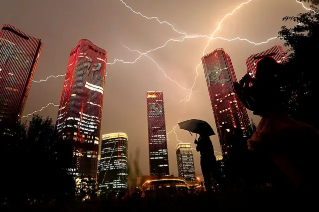 A bolt of lightning crosses the sky as people look at buildings displaying a light show on the eve of the 100th anniversary of the Chinese Communist Party in Beijing