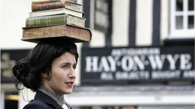 Una mujer con libros sobre su cabeza frente a un letrero de Hay-on-Wye